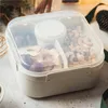 A beige lunchbox filled with nuts, dried fruit, and a small container, placed on a textured surface with some greenery and cinnamon sticks in the background. Podgląd zdjęcia produktu