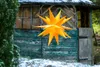 A yellow star-shaped decoration hangs in front of a weathered wooden shed in a snowy environment. Produktbild-Vorschau