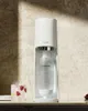 A white SodaStream soda maker is shown on a countertop next to a glass of sparkling water with raspberries. Produktbild-Vorschau