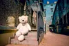A large white teddy bear sits on outdoor steps in front of a building covered in reflective blue tiles. Produktbild-Vorschau