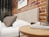 A close-up view of a bed with a beige upholstered headboard, white pillows, and a decorative brown pillow. The background features a brick wall and a framed poster with text. Produktbild-Vorschau