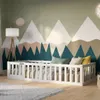 A white wooden bed frame with a gate is set up in a child's room with a mountain mural on the wall. Produktbild-Vorschau