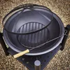 A black metal fire pit with a mesh cover and a poker tool, resting on a gravel surface. Produktbild-Vorschau