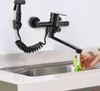 A black kitchen faucet with a sprayer is mounted on a wall above a sink, with a hand washing lettuce under the faucet. Náhled obrázku produktu