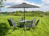 A garden table and chairs set with a large umbrella, set up on a grassy lawn with trees and a blue sky in the background. Produktbild-Vorschau