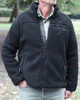 A man wearing a black fleece jacket and a green shirt stands outdoors in a park-like setting. Anteprima dell’immagine prodotto