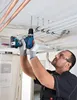 A man is using a cordless drill to drill into a ceiling with pipes and cables visible. Náhled obrázku produktu