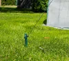 A close-up view of a green stake in the grass, with a white greenhouse in the background. Produktbild-Vorschau