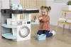 A young girl is playing with a toy laundry set, which includes a washing machine, ironing board, and accessories. Produktbild-Vorschau