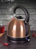 A copper-colored electric kettle is shown on a dark countertop, with steam rising from the spout. A glass of red liquid and some dried petals are also visible. Náhled obrázku produktu