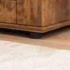 A close-up view of the bottom corner of a wooden cabinet, showing one of its black legs resting on a wooden floor next to a rug. Podgląd zdjęcia produktu