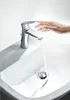 A hand is reaching towards a modern chrome faucet over a white sink, with water running. Podgląd zdjęcia produktu