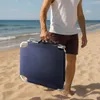 A person walking on a beach carrying a folded navy blue beach chair. Náhled obrázku produktu