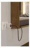 A white electrical outlet is shown on a wall, with a white power adapter plugged into it. A black power cord extends from the adapter and disappears behind a wooden shelf. The shelf has a built-in light that illuminates a pillow and bedding. Anteprima dell’immagine prodotto