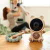 A baby monitor shaped like a bear is on a wooden table, with a baby teether next to it. In the background, a woman is holding a baby. Produktbild-Vorschau