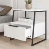 A white nightstand with a black metal frame is shown next to a bed. Podgląd zdjęcia produktu