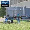 A blue and black log splitter is shown outdoors on a grassy lawn, with a building and fence in the background. Produktbild-Vorschau