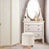 A white bouclé fabric ottoman is placed in front of a white dresser with a large oval mirror on top. A large white wardrobe is to the left of the dresser. Produktbild-Vorschau