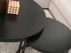 Two black nesting coffee tables are shown in a living room setting, with a blurred sofa in the background. Podgląd zdjęcia produktu