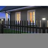 A black fence with solar lights in front of a brick house with illuminated windows at dusk. Voir les images du produit