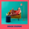 A man is relaxing on a yellow armchair, holding a remote control, with a floor lamp nearby. The background is teal, and the image has a pink border. Voir les images du produit