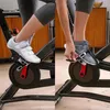 A split image showing a person's feet on an indoor cycling bike. The left side shows a white cycling shoe clipped into a pedal, and the right side shows a gray athletic shoe with a strap securing it to a pedal. Voir les images du produit