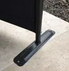 Close-up of the base of a black screen or divider, showing its metal foot and pole attached to a tiled patio. Produktbild-Vorschau