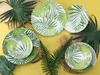 A collection of plates and bowls with a tropical leaf pattern, arranged on a yellow tablecloth with natural elements like wood and ferns. Náhľad obrázku produktu