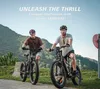 Two men riding electric mountain bikes on a paved road with a scenic mountain backdrop. Produktbild-Vorschau