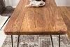 A close-up view of a wooden dining table with dark metal legs, placed on a patterned rug. A chair is partially visible on the left. Anteprima dell’immagine prodotto