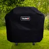 A black gas grill cover with the TAINO logo is shown outdoors on a grassy lawn with trees and plants in the background. Produktbild-Vorschau