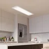 A modern kitchen with white cabinets, a stainless steel refrigerator, and a long, rectangular LED panel light hanging from the ceiling. Produktbild-Vorschau