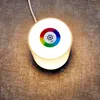 A top-down view of a white LED table lamp with a color-changing ring on its surface, placed on a textured brown and gray surface. Produktbild-Vorschau