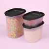 Three clear plastic food storage containers with black lids, two stacked on the right and one larger one on the left. The largest container is filled with colorful cereal loops, the top container on the right is filled with gummy candies, and the bottom co Anteprima dell’immagine prodotto