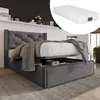 A dark gray upholstered bed frame with a tufted headboard and a mattress is shown in a room setting. The bed has storage drawers underneath, and a blanket is draped over the mattress. Produktbild-Vorschau