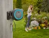 A woman is watering flowers in a garden with a hose reel mounted on a wall. Voir les images du produit