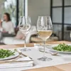 Two wine glasses are on a table, one filled with white wine, next to a plate of salad. A person is visible in the background. Anteprima dell’immagine prodotto