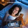 A woman is lying in bed looking at her phone, which displays a live feed from a baby monitor camera showing a sleeping baby. Produktbild-Vorschau