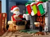 A LEGO Santa Claus figure sitting in a chair, holding a cookie and a mug, with Christmas stockings hanging above a fireplace. Podgląd zdjęcia produktu