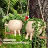 Two cute, smiling plant pots shaped like little characters are hanging from a tree branch by ropes, with plants spilling out of them. Anteprima dell’immagine prodotto