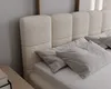 A close-up view of a bed with a textured cream-colored headboard and light gray pillows. Náhled obrázku produktu