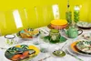 A table setting with tropical-themed tableware, including plates, cups, a teapot, and bottles, arranged on a white tablecloth. Anteprima dell’immagine prodotto
