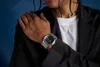 A person wearing a black blazer and a white t-shirt, showing off a silver G-SHOCK watch on their wrist. Náhled obrázku produktu