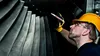 A man in a yellow hard hat and safety glasses inspects a large industrial fan or turbine with a marker. Produktbild-Vorschau