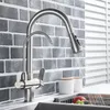 A kitchen faucet with a pull-out spray head is shown in use, with water flowing into a sink containing fruit. The background features a gray brick tile backsplash and white blinds. Náhľad obrázku produktu