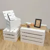 Two stacks of white wooden crates are displayed against a light gray wall and a wooden floor. One stack has an open crate on top, revealing a black object inside. The other stack has decorative items on top, including a candle, dried lavender, and a framed Produktbild-Vorschau