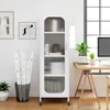 A white, narrow, five-tier cabinet with glass doors and gold handles stands in a room with artwork on the wall and a desk with plants and a computer. Podgląd zdjęcia produktu