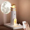 A lighthouse-shaped night light is placed on a book next to a moon projection lamp. Produktbild-Vorschau