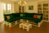 A living room with a sectional sofa made of pallets and green cushions, a coffee table with drinks, and bookshelves. Anteprima dell’immagine prodotto