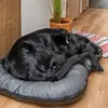 A black dog is sleeping on a gray dog bed on a wooden floor. A plant in a basket and a striped rug are in the background. Produktbild-Vorschau